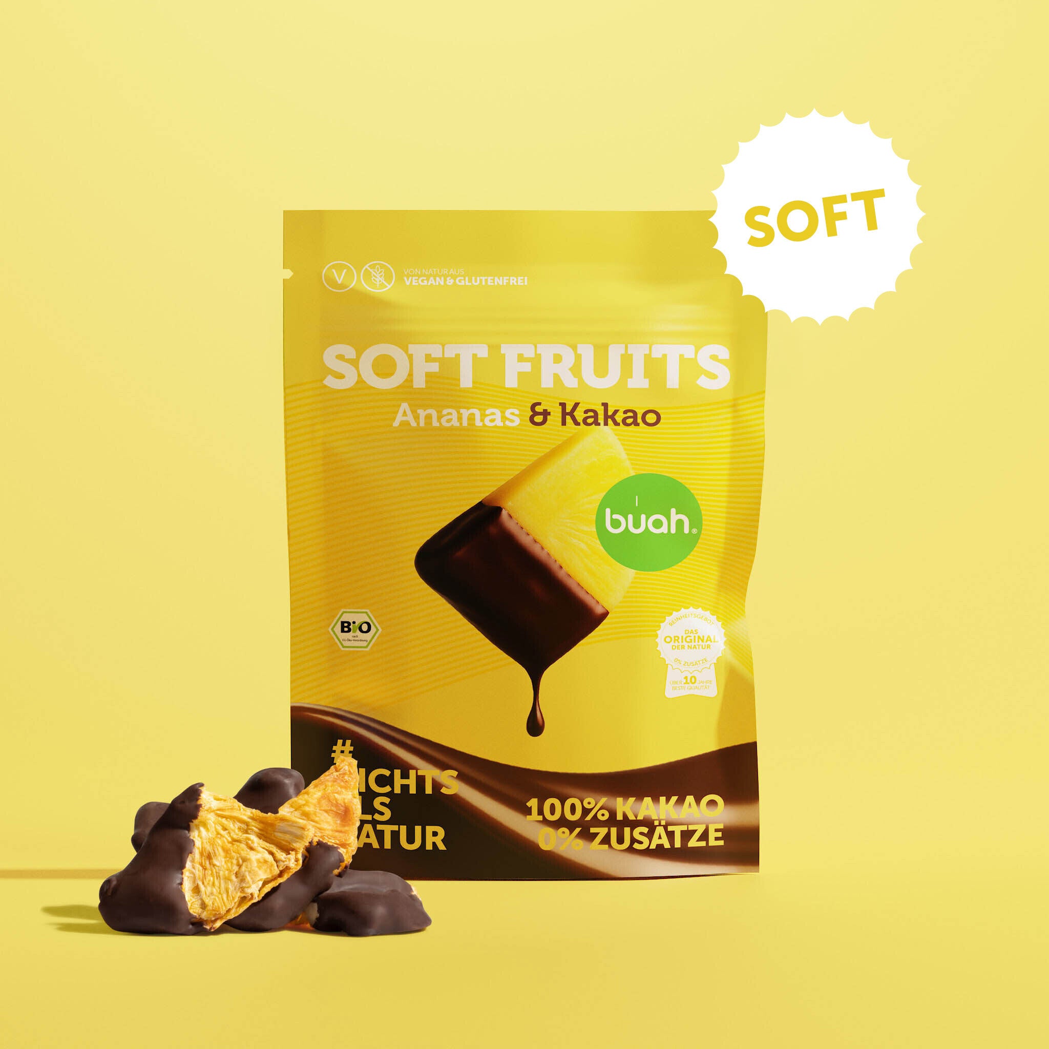 Ein gelber Beutel buah Soft Fruits der Sorte Ananas & Kakao steht vor einem gelben Hintergrund. Davor liegen einige softe, mit Schokolade überzogene Ananas-Stücke.