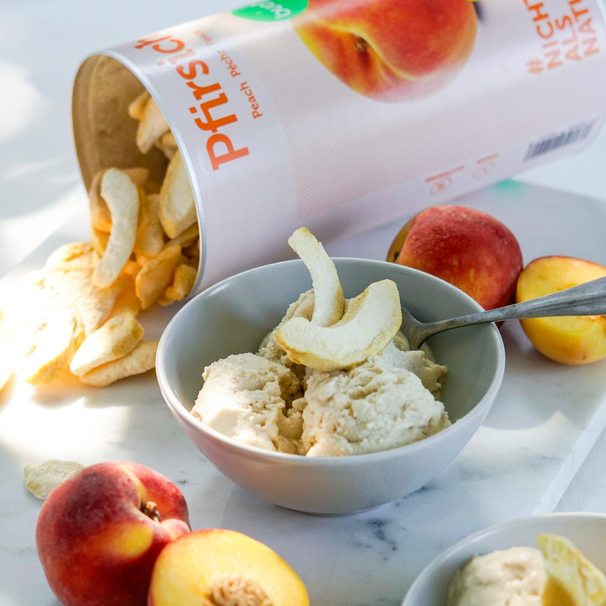 Eine graue Schale mit Eiscreme ist mit knusprigen PfirsichstĂĽcken garniert. Dahinter liegt eine umgekippte groĂźe Dose buah gefriergetrocknete FrĂĽchte der Sorte Pfirsich, aus der weitere FruchtstĂĽcke auf eine helle Marmorplatte rieseln. Zur Dekoration liegen frische Pfirsiche daneben.