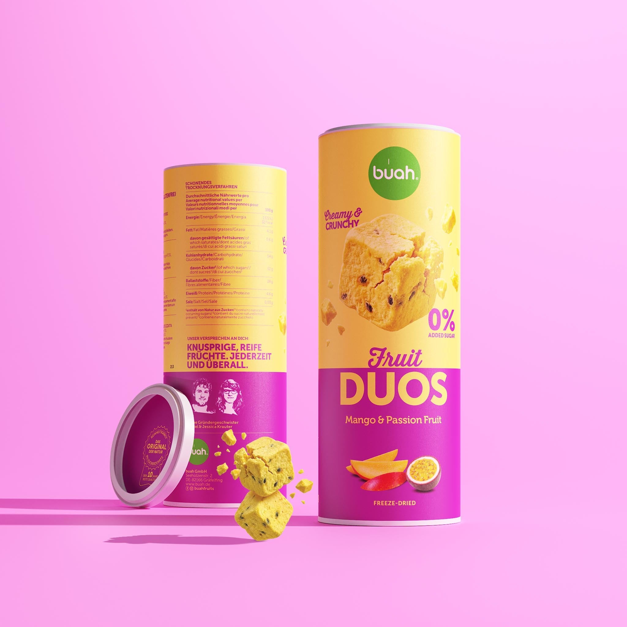 Eine Slim-Dose buah Fruit DUOS Mango & Passion Fruit steht vor einem rosafarbenen Hintergrund. Dahinter steht eine zweite Dose, die ihre Rückseite zeigt. Der Deckel lehnt seitlich an. Vorne schweben ein paar gelbe Fruchtwürfel in der Luft.