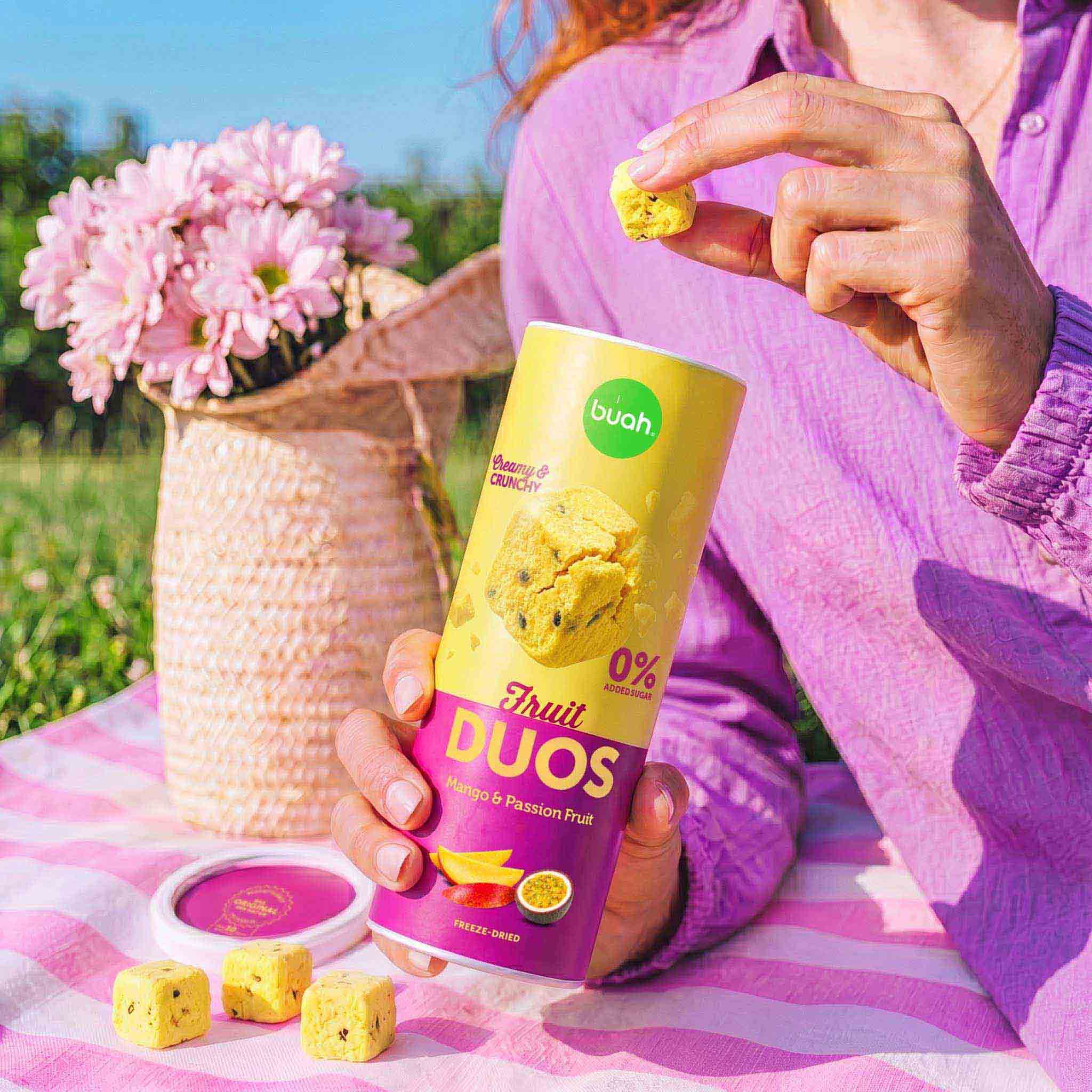 Eine Person sitzt auf einer Picknickdecke im Gras. Sie hält ein quadratisches Stück buah Fruit DUOS Mango & Maracuja zwischen Daumen und Zeigefinger. In der anderen Hand hält sie die geöffnete Slim-Dose, vor ihr liegen weitere Fruchtwürfel.