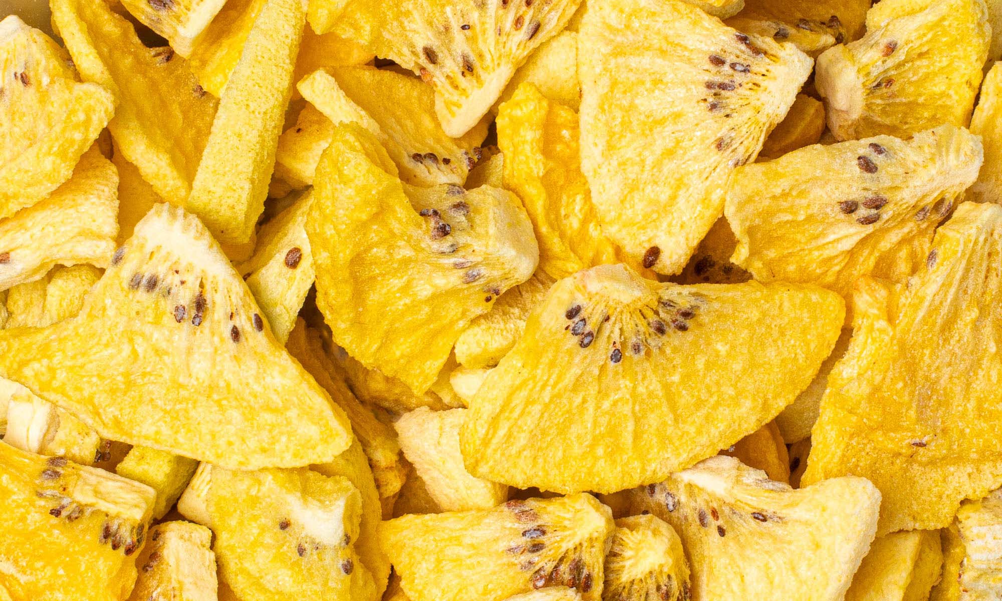 Eine formatfüllende Nahaufnahme von vielen texturierten, buah gefriergetrockneten Kiwi-Stücken. Man erkennt deutlich die gelbe Farbe und die kleinen schwarzen Kerne.