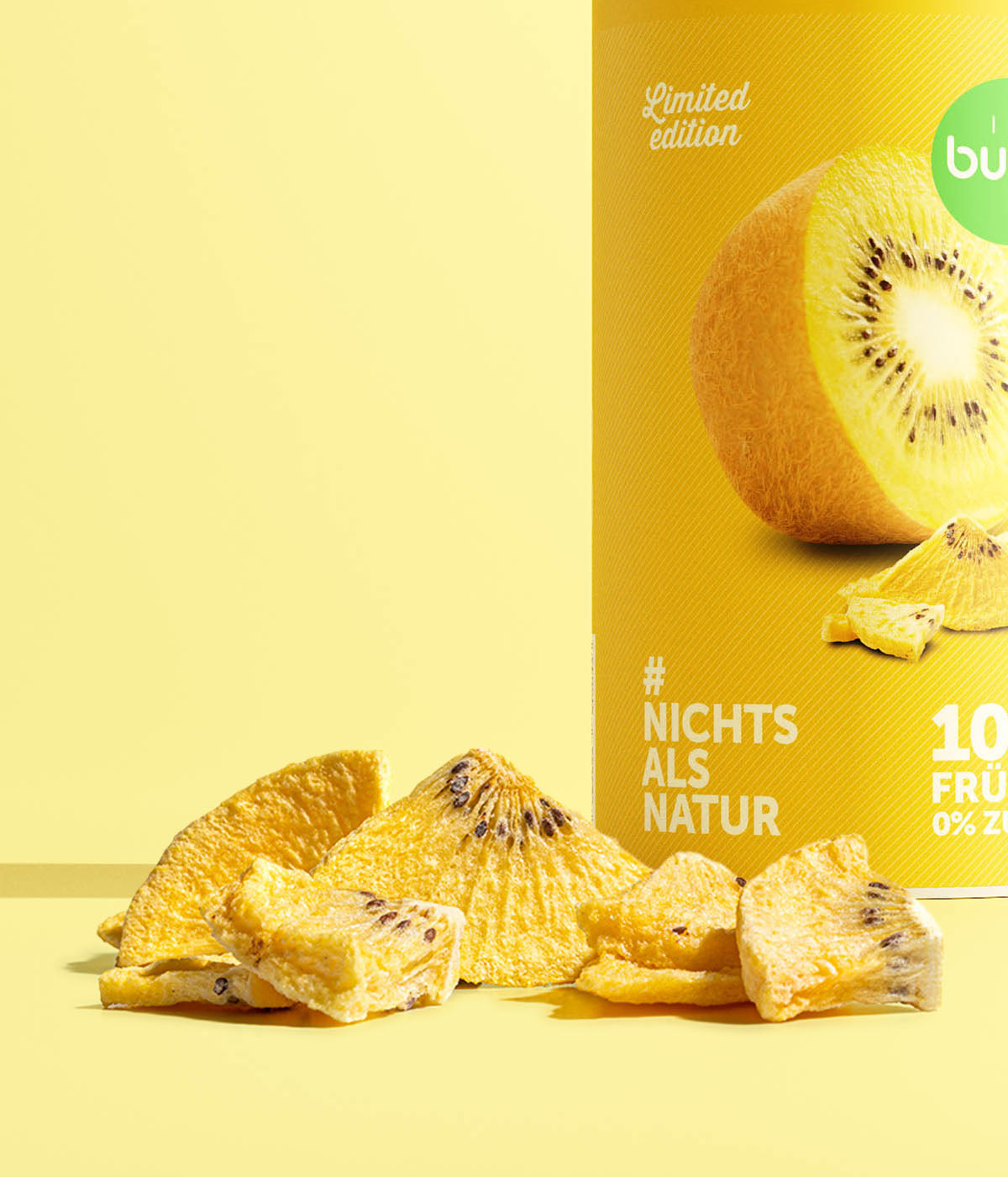 Eine Nahaufnahme der gelben Dose buah gefriergetrocknete Goldene Kiwi. Im scharf fokussierten Vordergrund liegen knusprige, hellgelbe Kiwi-Stücke mit schwarzen Kernen auf einem gelben Hintergrund.
