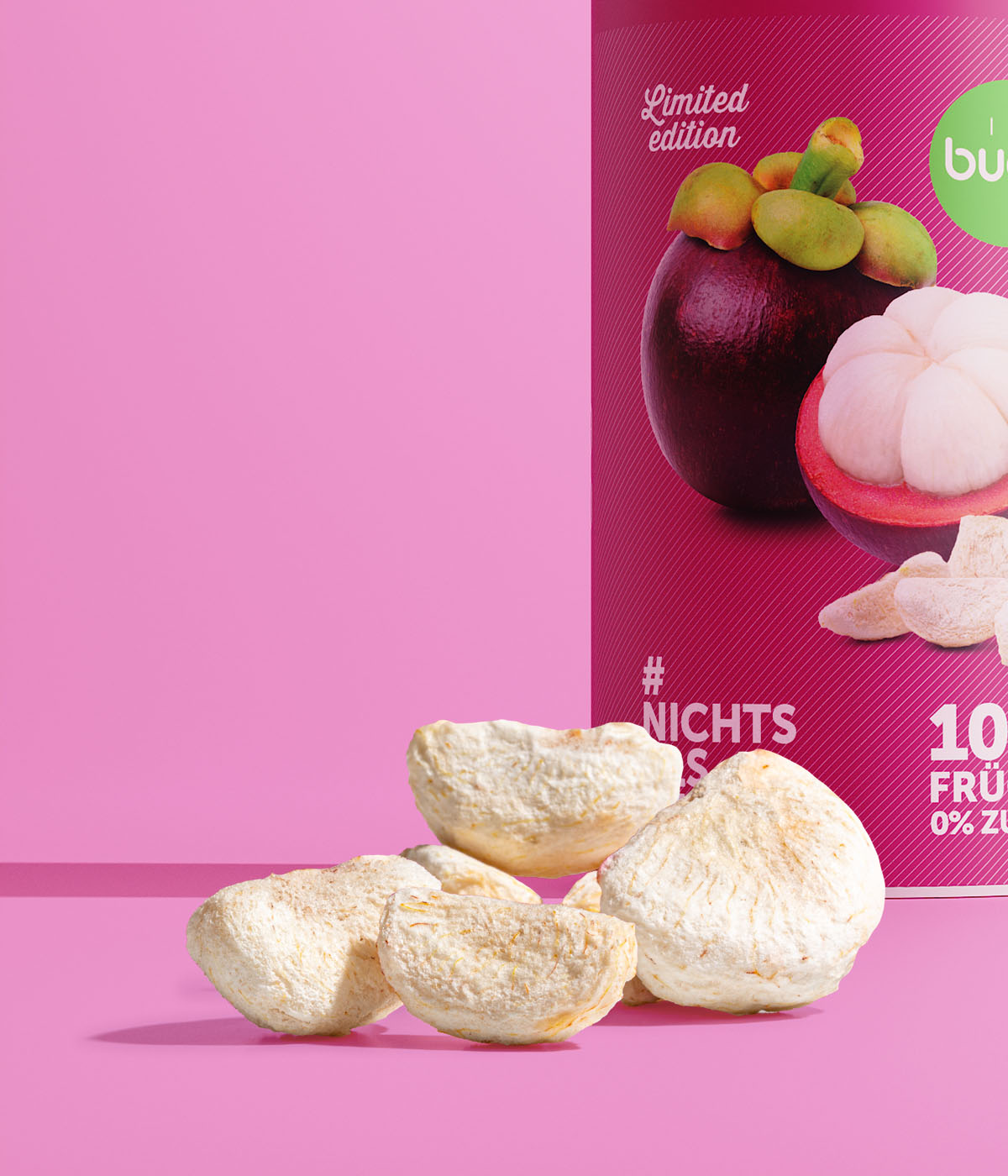 Eine Nahaufnahme von buah gefriergetrockneten Mangostan-StĂĽcken im Vordergrund. Dahinter steht eine angeschnittene Dose der Limited Edition Mangostan vor einem pinken Hintergrund.