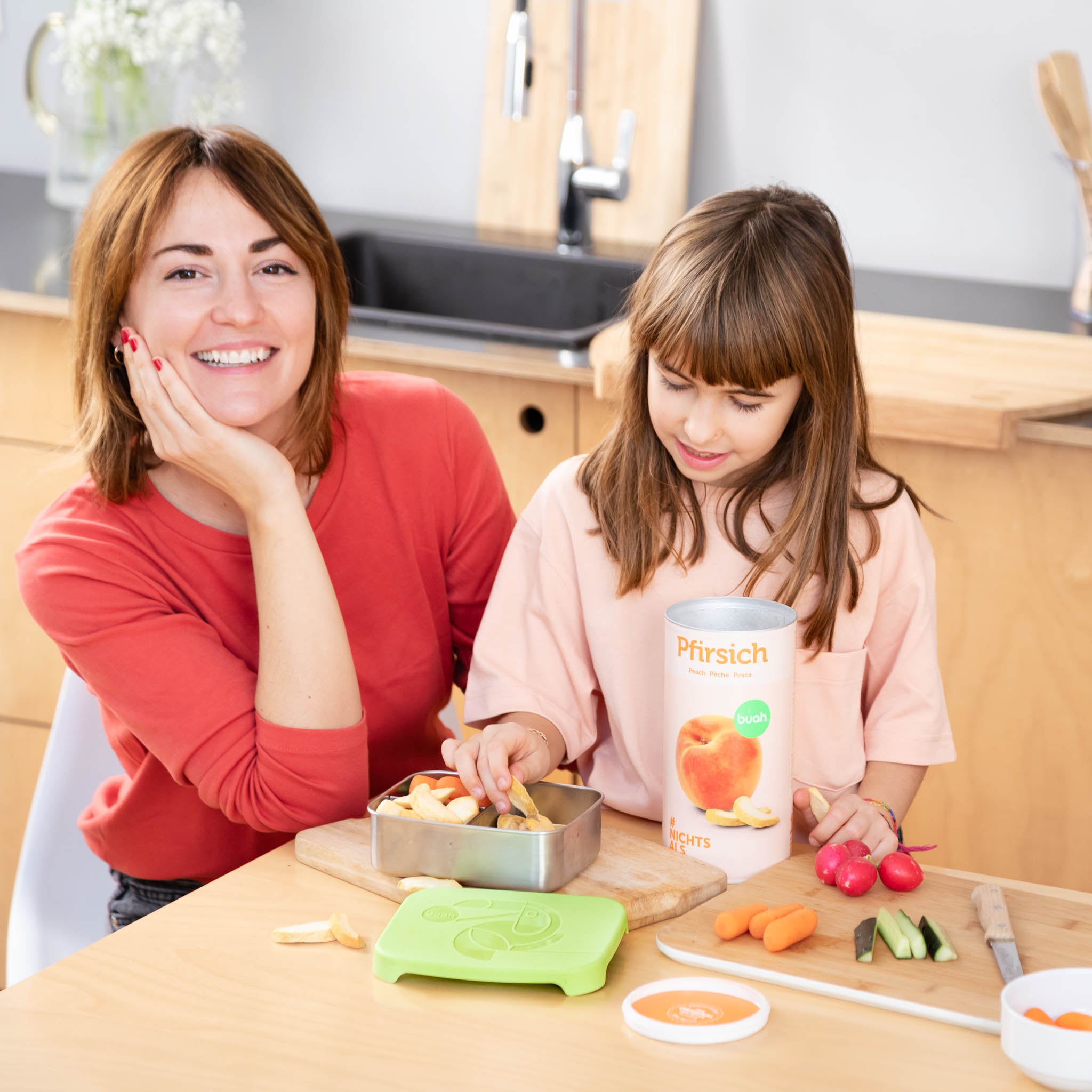 Mama und Tochter sitzen am Kuechentisch und essen buah gefriergetrockneten Pfirsich aus der buah Lunchbox. 
