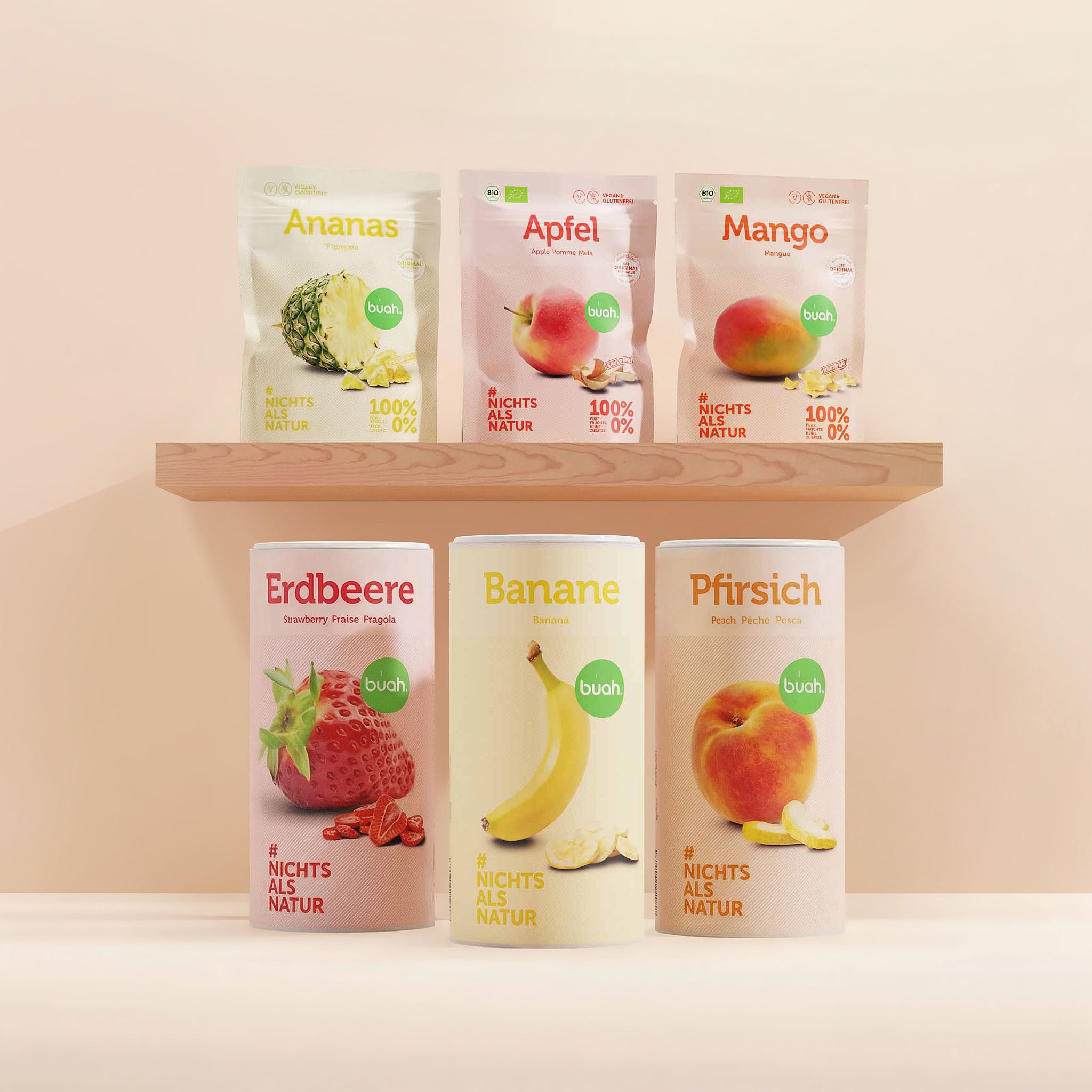 buah Kids-Snack-Set. Ein Regal aus hellem Holz mit drei Snackpacks gefriergetrocknete Früchte der Sorten Ananas, Apfel und Mango. Darunter stehen auf dem Boden drei große Dosen der Sorten Erdbeere, Banane und Pfirsich vor einem beigefarbenen Hintergrund.