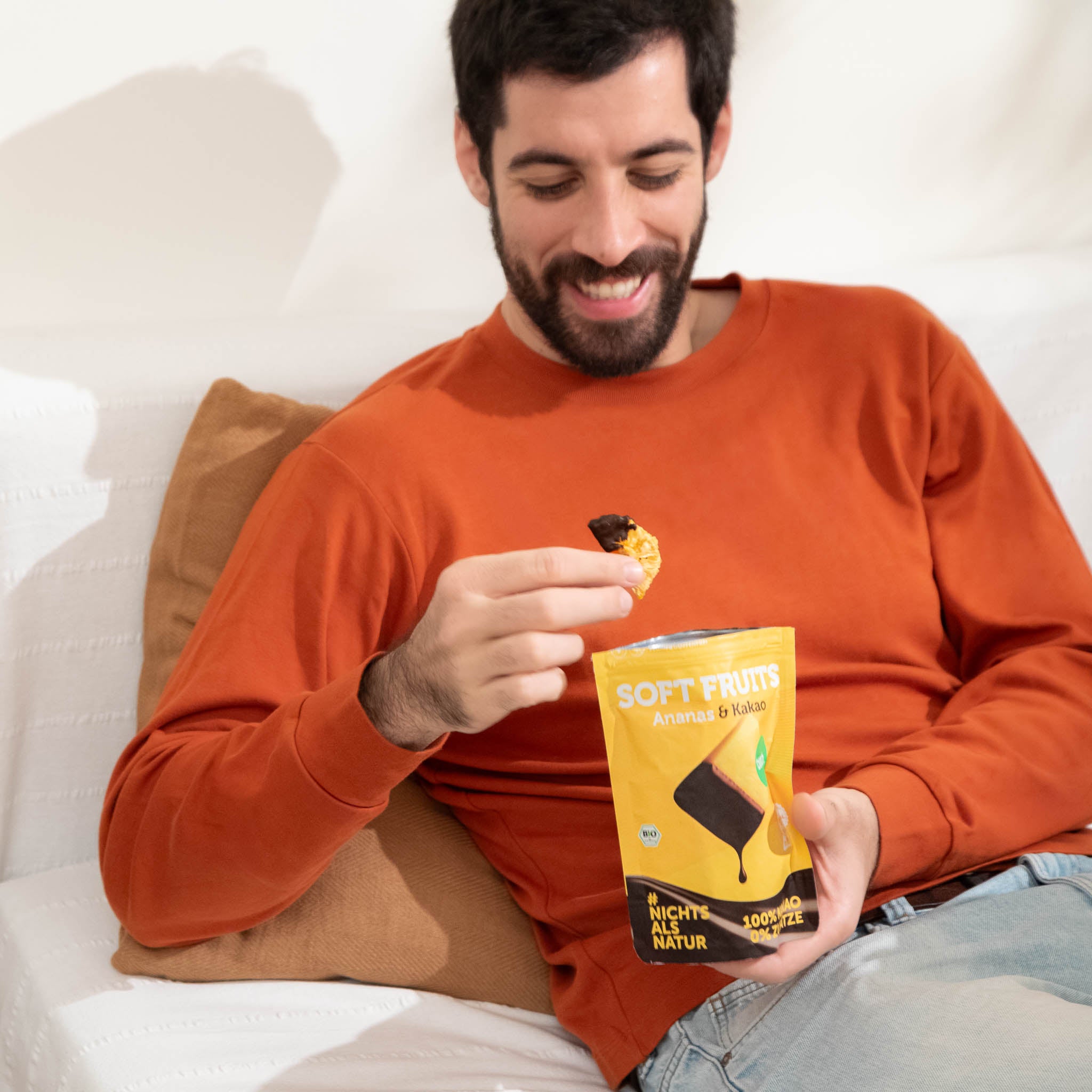 Ein lächelnder Mann im orangefarbenen Pullover sitzt gemütlich auf einem weißen Sofa und isst einen schokolierten Fruchtsnack. In der anderen Hand hält er einen geöffneten gelben Beutel buah Soft Fruits der Sorte Ananas & Kakao.