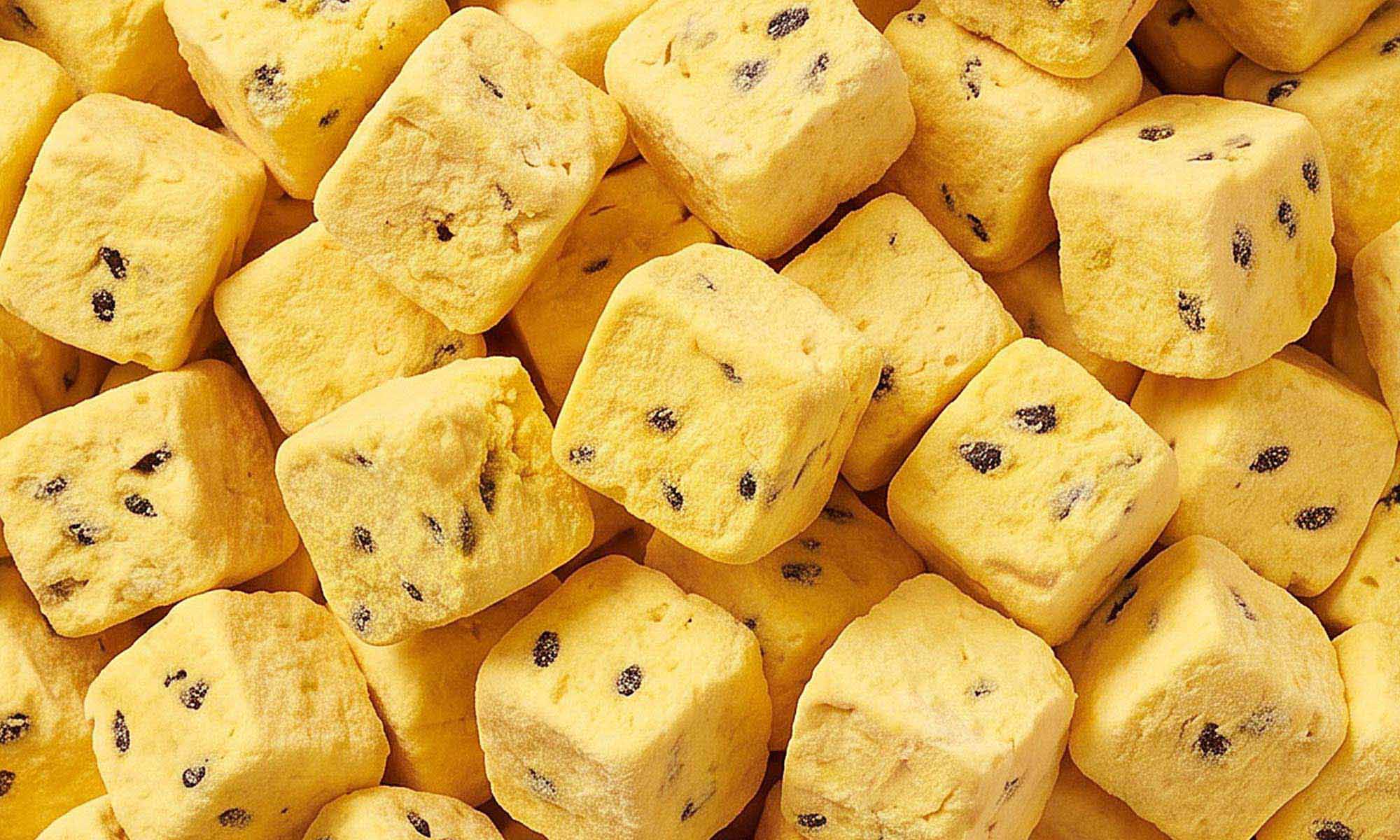 Eine formatfüllende Nahaufnahme von vielen quadratischen, buah gefriergetrockneten Fruchtwürfeln der Sorte Mango & Maracuja. Die gelben Würfel sind mit dunklen Kernen durchzogen.