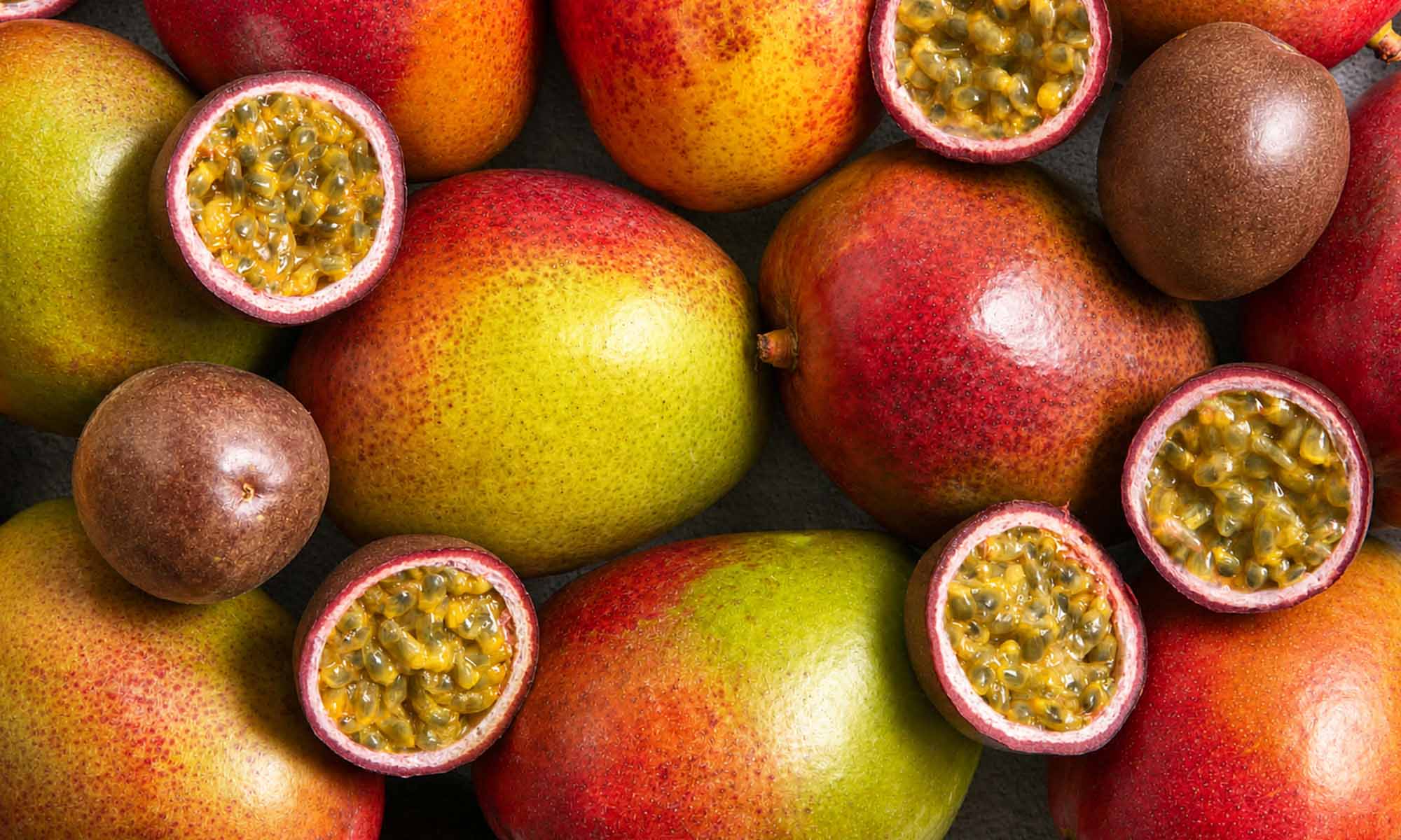 Eine farbenfrohe Nahaufnahme von vielen frischen, ganzen Mangos. Dazwischen liegen ganze und aufgeschnittene Maracujas, die ihr saftiges, von Kernen durchzogenes Fruchtfleisch zeigen.