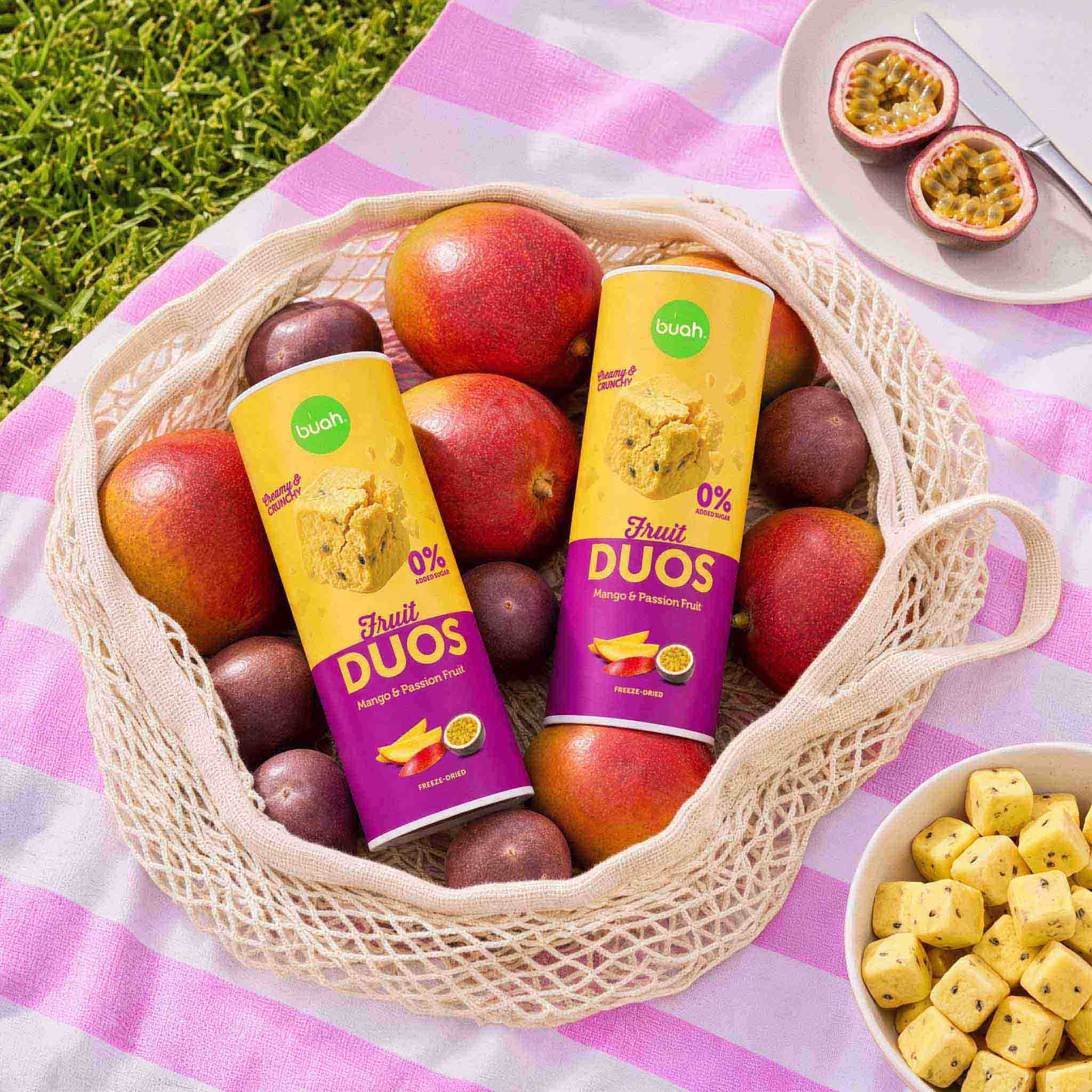 Ein Einkaufsnetz voller frischer Mangos und Maracujas liegt auf einer Decke im Gras. Mittendrin stecken zwei Slim-Dosen buah Fruit DUOS Mango & Maracuja. Daneben steht eine Schale voller gefriergetrockneter Fruchtwürfel.