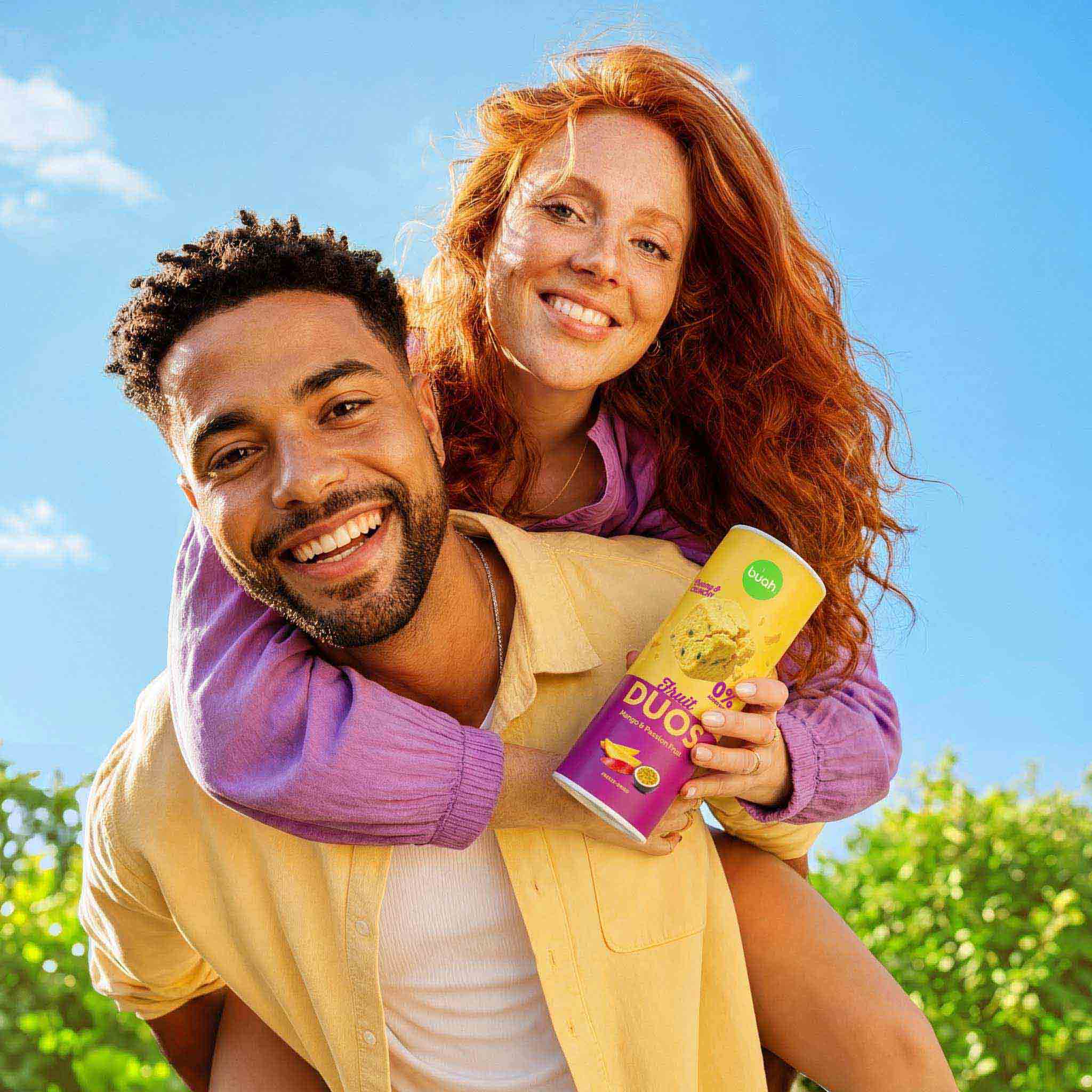 Ein fröhliches Paar an einem sonnigen Tag. Ein Mann trägt eine rothaarige, lächelnde Frau huckepack. Sie hält eine hohe Slim-Dose buah Fruit DUOS Mango & Passion Fruit in der Hand.