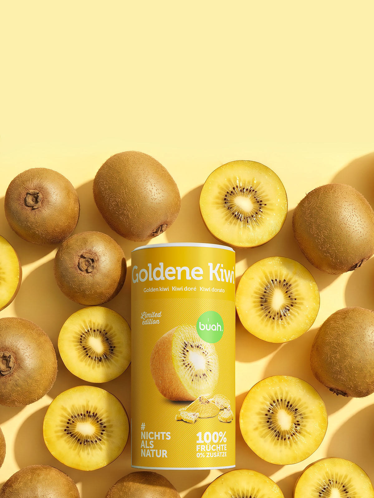 Eine flach von oben fotografierte Anordnung im Hochformat auf hellgelbem Hintergrund. Im Zentrum steht eine gelbe Dose buah gefriergetrocknete Goldene Kiwi (Limited Edition), umgeben von zahlreichen frischen, ganzen und halbierten Goldenen Kiwis.