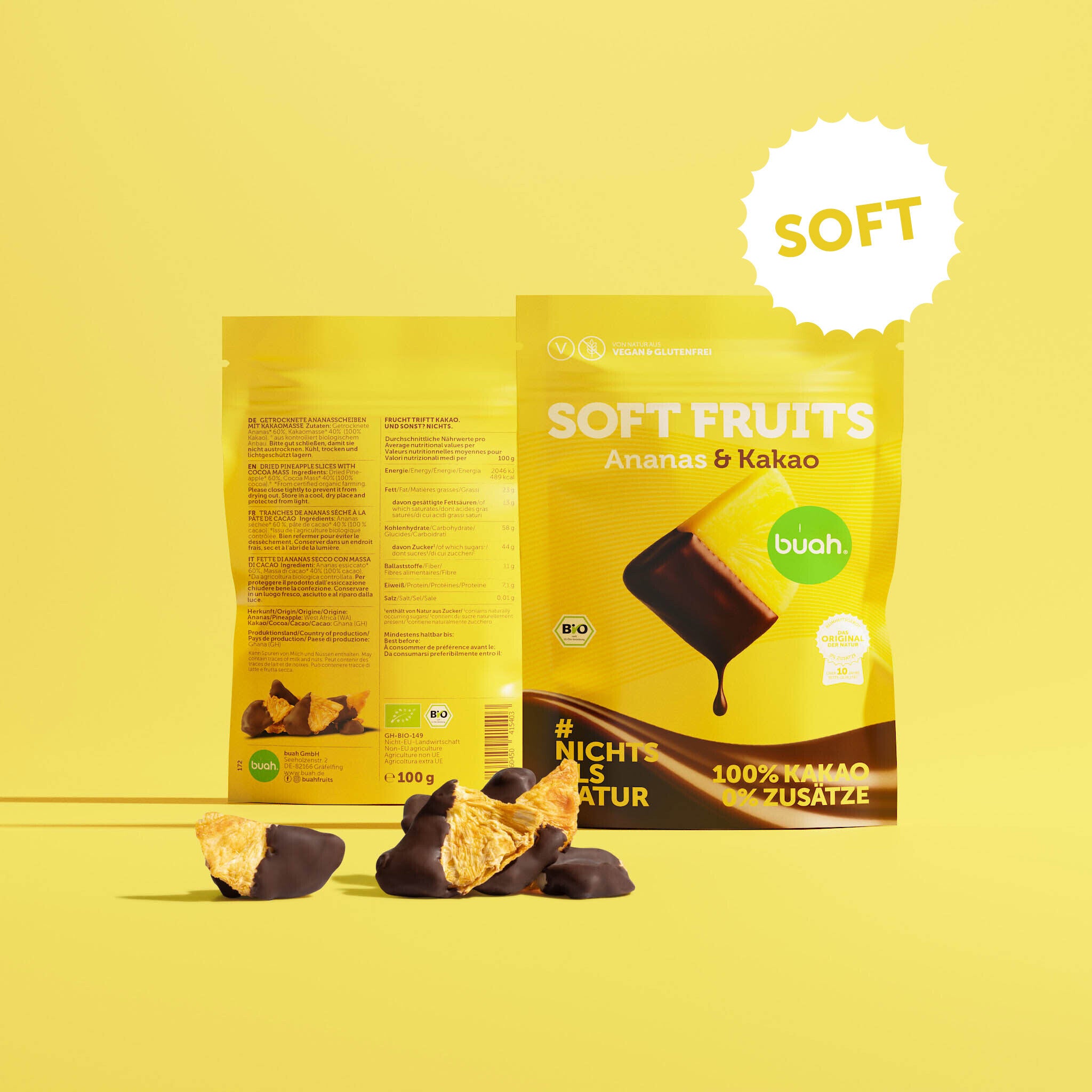Ein gelber Beutel buah Soft Fruits der Sorte Ananas & Kakao steht vor einem gelben Hintergrund. Dahinter ist die Rückseite der Verpackung zu erkennen. Davor liegen einige softe, mit Schokolade überzogene Ananas-Stücke.